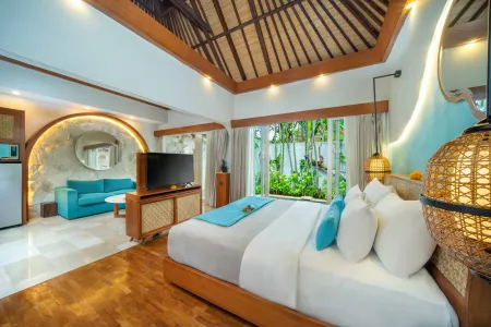 Luxurious Balinese-style bedroom suite with king bed, blue sofa, TV, and tropical garden views through arched windows.