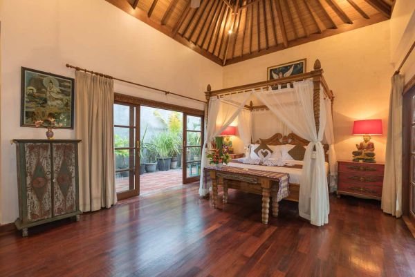 Elegant Balinese villa bedroom with four-poster canopy bed, wooden accents, red lamps, and open doors to lush garden.