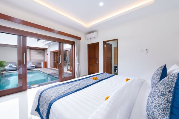 Luxurious Balinese villa bedroom with white king bed, blue bedding, and floor-to-ceiling doors opening to a private turquoise pool.