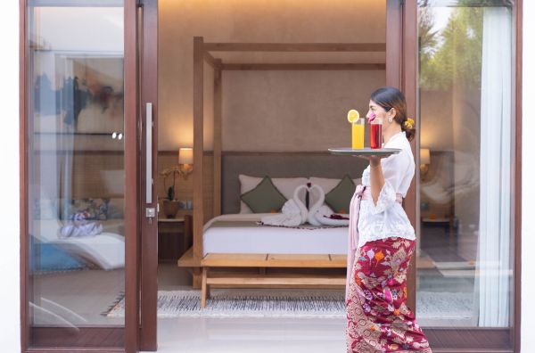 Balinese woman in traditional sarong serving fresh orange juice on a tray in a luxurious tropical bedroom with four-poster bed and open balcony doors.