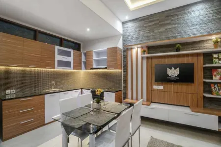 Modern wooden kitchen with L-shaped counters, glass dining table, white chairs, and built-in TV entertainment unit in a bright, open interior. - Bali Villas
