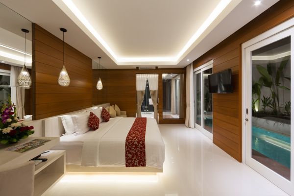 Luxurious Bali-style bedroom with wooden walls, white king bed with red accents, balcony doors to infinity pool, and tropical plants.