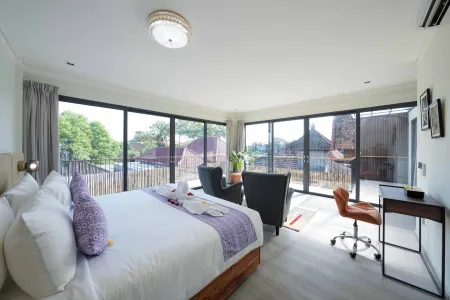 Spacious modern bedroom with king bed, purple accents, desk, seating area, and panoramic tropical views through large windows.
