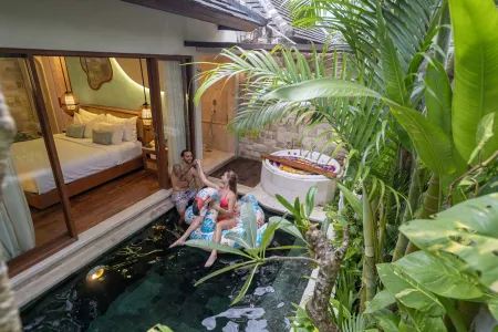 Romantic couple relaxing on an inflatable float in a private plunge pool at a luxurious Bali-style villa, surrounded by lush tropical plants and an open indoor bedroom.