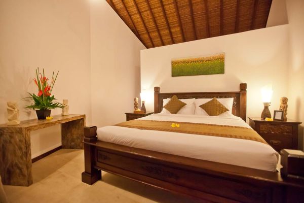 Spacious Balinese bedroom with carved wooden four-poster bed, tropical flower vase, and green field artwork on white walls.