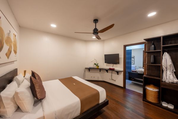 Spacious modern bedroom with king bed, wooden ceiling fan, wall-mounted TV, abstract wall art, and open door to en-suite bathroom.