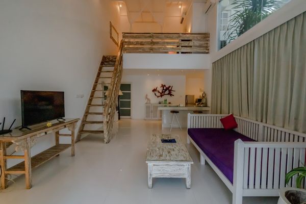 Spacious Balinese loft living room with wooden mezzanine stairs, purple sofa, TV console, white coffee table, and tropical wall art. - Bali Villas