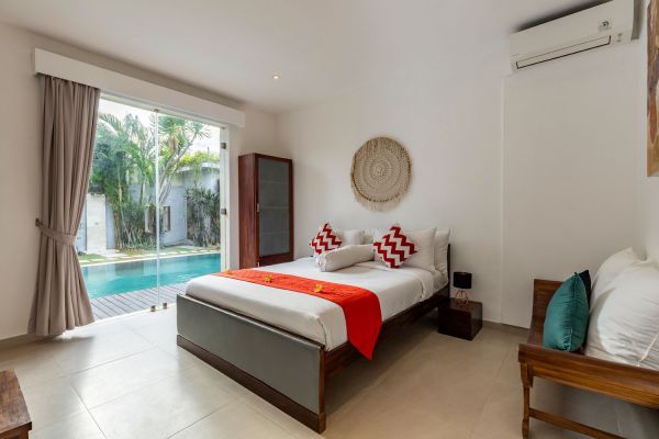 Spacious Bali villa bedroom with king bed, red accents, and sliding doors opening to private pool and tropical garden.