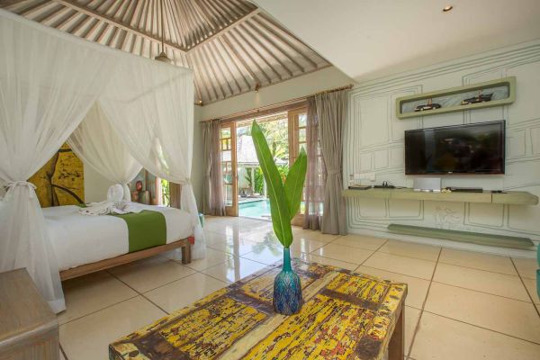 Spacious Balinese villa bedroom with white canopy bed, open doors to infinity pool, tropical plant accents, and wooden furniture.