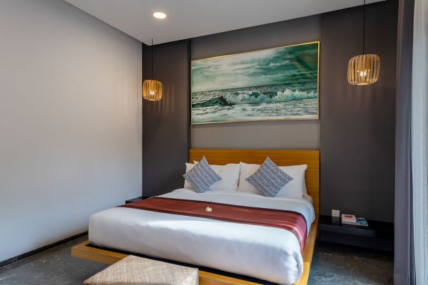 Modern tropical bedroom featuring a low wooden platform bed with white linens, red throw, gray pillows, abstract ocean wave painting, and rattan pendant lights.