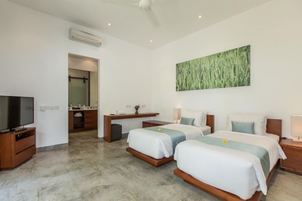 Spacious Balinese villa bedroom with twin wooden beds, teal accents, rice field wall art, TV, and open en-suite bathroom.