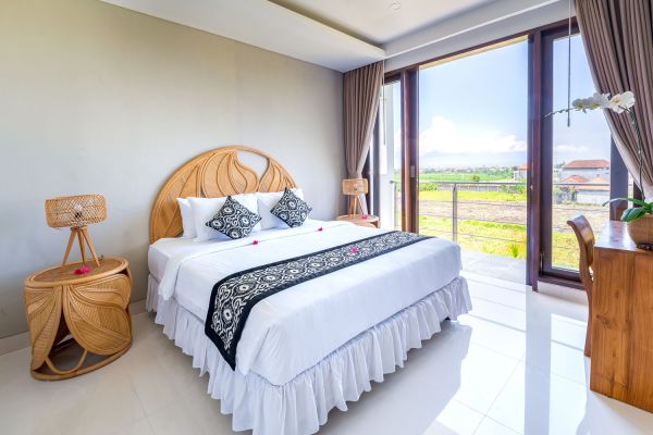Serene Balinese bedroom with ornate wooden headboard, crisp white bedding, rattan lamps, and tropical rice field view through open balcony doors.