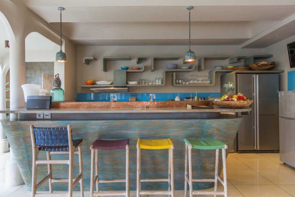 Rustic tropical kitchen bar with copper counter, colorful woven stools, wooden shelves stocked with dishes, and stainless steel fridge. - Bali Villas