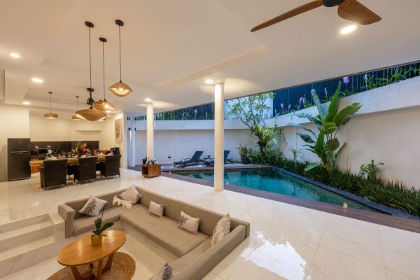 Modern Balinese villa interior featuring open-plan kitchen, lounge with gray sofa, and adjacent tropical pool garden. - Bali Villas