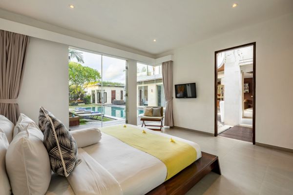 Spacious tropical bedroom in Bali villa with wooden bed, yellow throw, and floor-to-ceiling doors opening to infinity pool and palm garden.