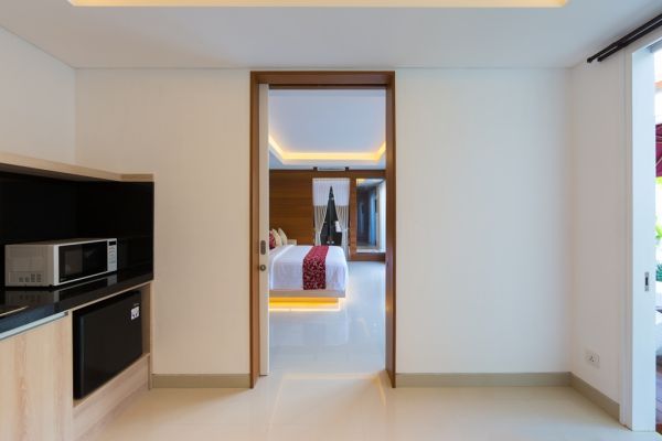Modern Balinese hotel suite interior with kitchenette, microwave, mini-fridge, and open door to bedroom with red-bedded king bed.