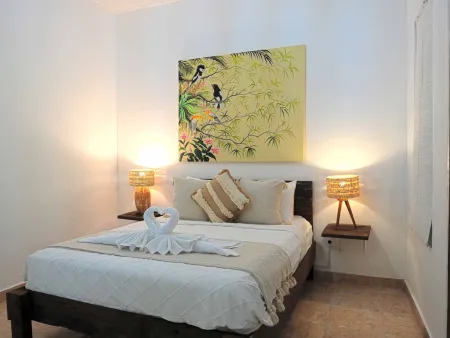 Cozy tropical bedroom with wooden bed, white linens, towel swan, rattan lamps, and vibrant bird-and-leaf wall art.