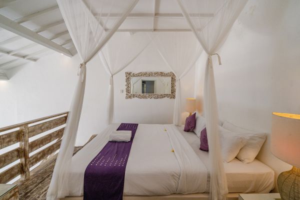 Elegant white canopy bed with purple runner and pillows in a airy loft bedroom with wooden railing and ornate mirror.