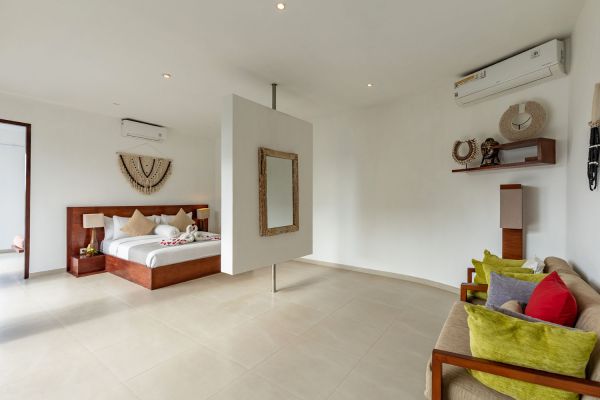 Luxurious tropical bedroom in a Bali villa featuring a king bed with rose petals, suspended wall mirror, woven decor, and plush green sofa.