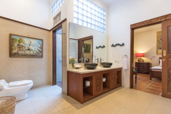 Spacious Balinese-style ensuite bathroom featuring wooden double vanity with black sinks, tropical artwork, and open doorway to cozy bedroom.