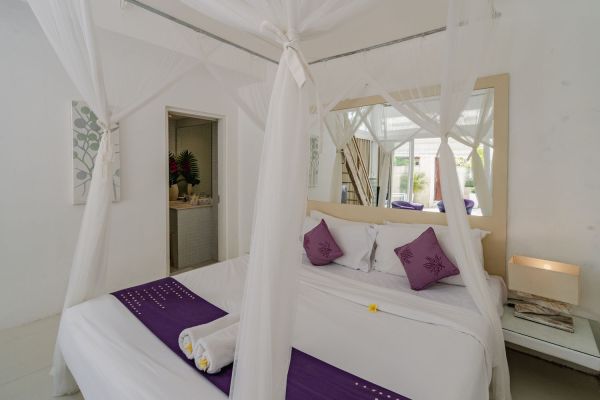 Elegant Balinese bedroom with white canopy bed, purple pillows, and mirrored vanity in airy tropical setting.