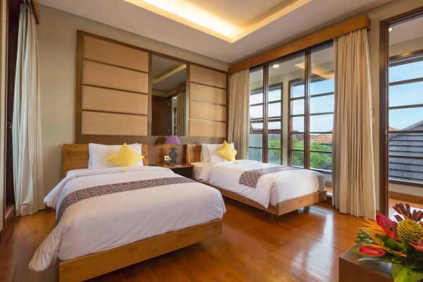 Cozy Balinese twin bedroom with wooden beds, yellow pillows, large window views of greenery, and tropical flower vase.