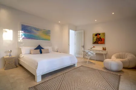 Bright, modern bedroom with white king bed, colorful abstract wall art, open door, stylish desk corner, and woven diamond-pattern rug.
