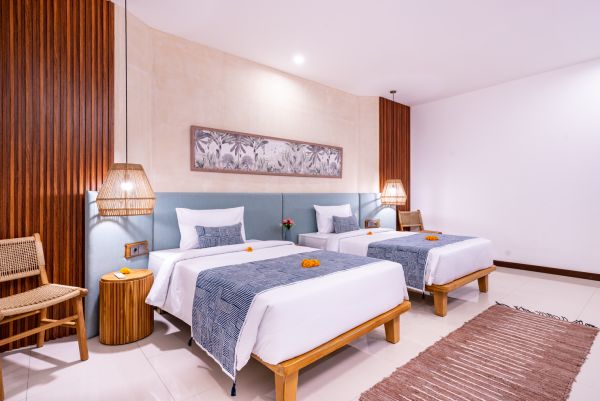 Cozy Balinese-style twin bedroom with wooden beds, blue batik textiles, rattan lamps, and tropical wall art.