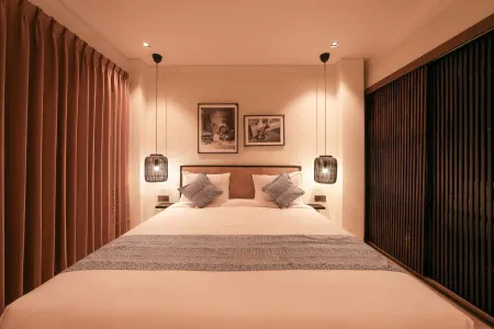 Modern minimalist hotel bedroom with a king bed, pendant lamps, framed black-and-white photos, maroon curtains, and wooden slatted walls.