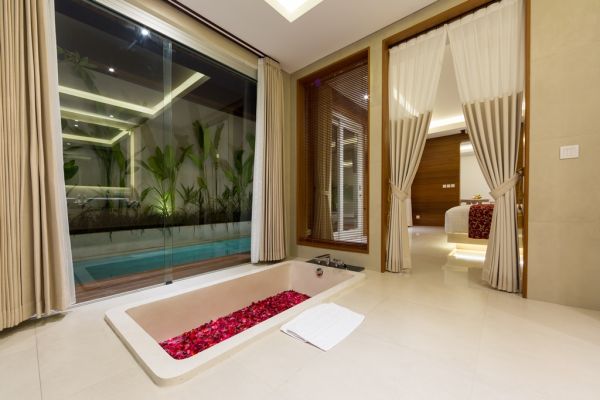 Luxurious modern bathroom with rose petal-filled bathtub, large windows overlooking nighttime infinity pool and tropical garden, and open doorway to bedroom.