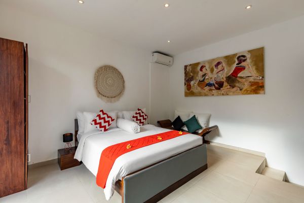Cozy Balinese villa bedroom featuring a king-size bed with red zigzag pillows and runner, wooden furniture, rattan wall decor, and vibrant wall art.
