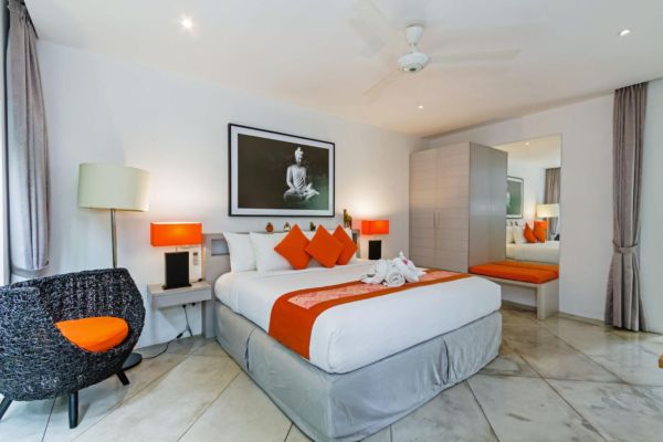 Luxurious Balinese hotel bedroom with king bed in orange accents, Buddha wall art, rattan chair, and ceiling fan.