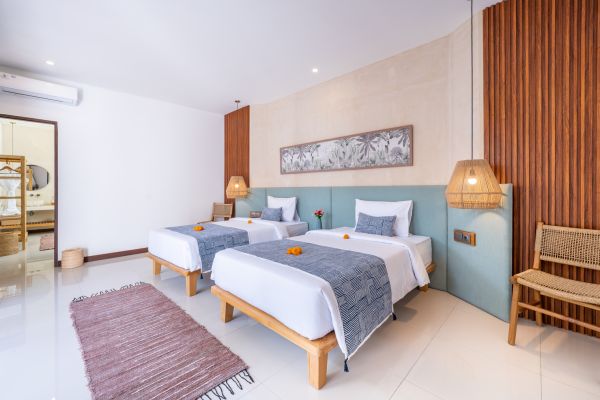 Spacious Balinese twin bedroom with wooden beds, blue textile accents, rattan lamps, and warm wood paneling.