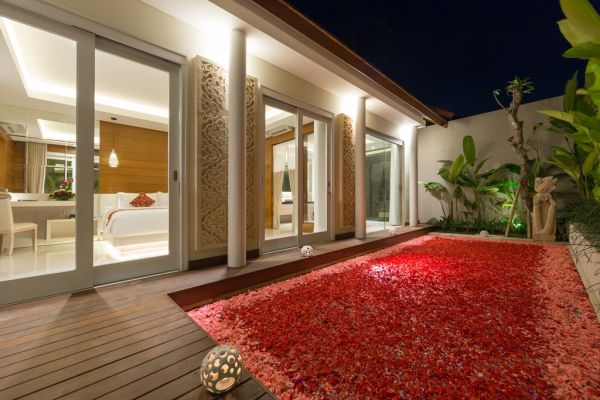 Luxurious Bali villa terrace at night with open sliding doors to a bedroom, overlooking a red petal-covered pool surrounded by tropical plants, statues, and glowing lanterns.