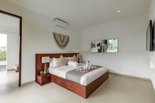 Spacious modern Balinese bedroom with wooden platform bed adorned with rose petal swan, white walls, woven wall hanging, and balcony access.