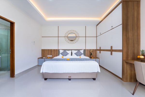 Modern Bali villa bedroom with white king bed, wooden accents, round mirror, tropical flowers, and glass shower door.