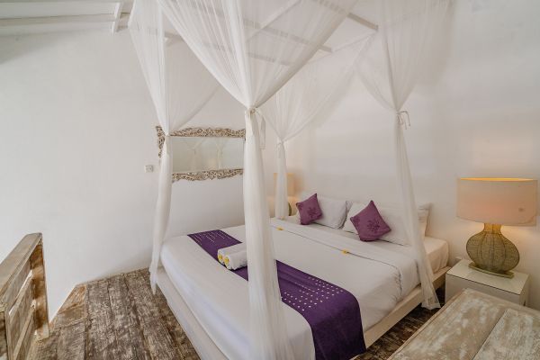 Cozy Balinese bedroom with white canopy bed, purple pillows and runner, folded towels, and wooden accents. - Bali Villas