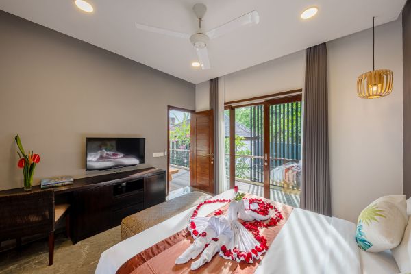 Luxurious tropical bedroom with heart-shaped rose petal decor on king bed, sliding doors to lush balcony, TV, and warm ceiling fan lighting. - Bali Villas
