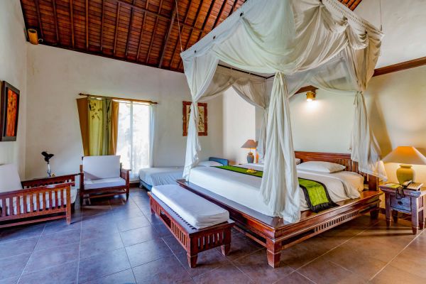 Spacious Balinese-style bedroom featuring a white-canopied king bed with green linens, wooden furniture, and tropical artwork.