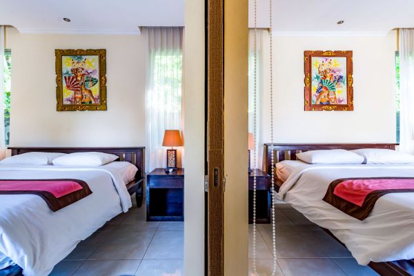 Elegant Balinese-style bedroom with a king bed, white linens, red throw, wooden nightstands, colorful framed art, and sheer curtains, viewed through open sliding doors.