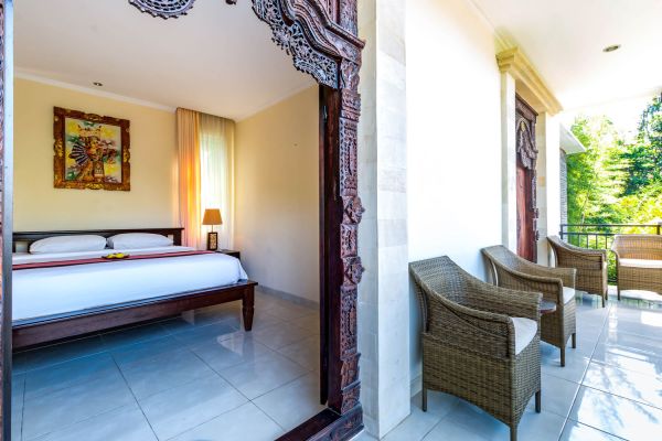Luxurious Balinese bedroom with carved wooden bed and artwork, opening to private balcony with rattan chairs surrounded by tropical greenery.