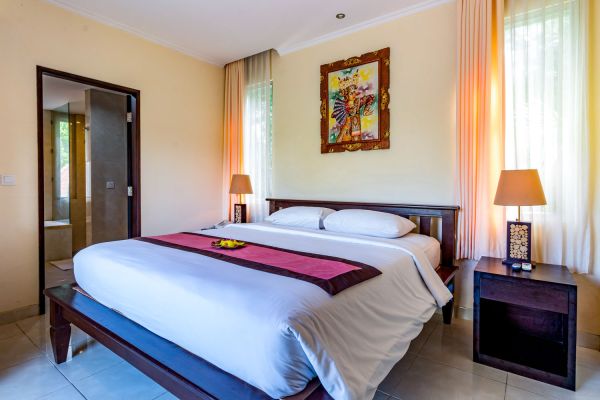 Spacious Balinese bedroom with a king-sized white bed, wooden accents, tropical artwork, and sheer orange curtains.