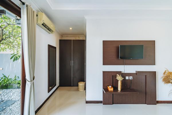 Modern Balinese hotel bedroom with wooden wardrobe, mounted TV, air conditioner, tropical garden view, and neutral decor.
