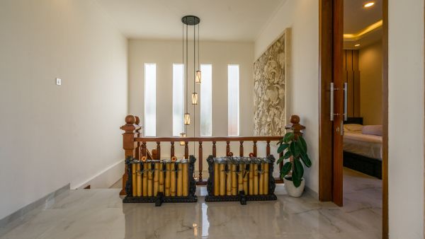 Modern tropical interior with wooden staircase balustrade, hanging pendant lights, potted plants, floral wall art, and two cushioned chairs on tiled floor leading to bedroom.