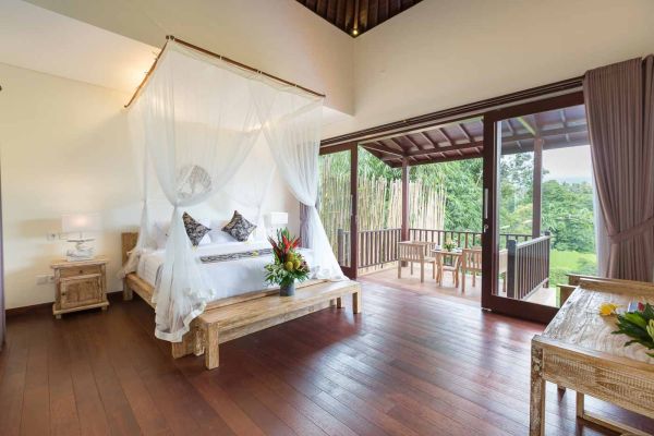 Luxurious Balinese bedroom featuring a white canopy bed, wooden floors, and sliding doors to a balcony overlooking lush tropical greenery.