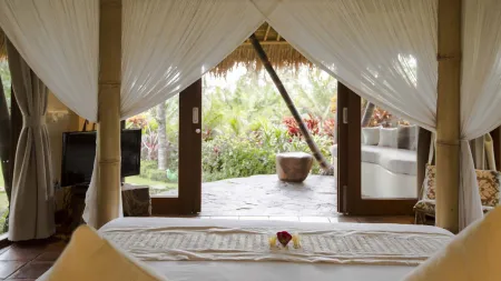 Luxurious Bali-style bedroom with a white-draped king bed and frangipani accents, opening via bamboo-framed doors with sheer curtains to a lush tropical garden.