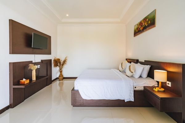 Modern minimalist bedroom with a wooden platform bed, white linens, wall-mounted TV, bedside lamps, and tropical decor like pampas grass and floral artwork on white walls.