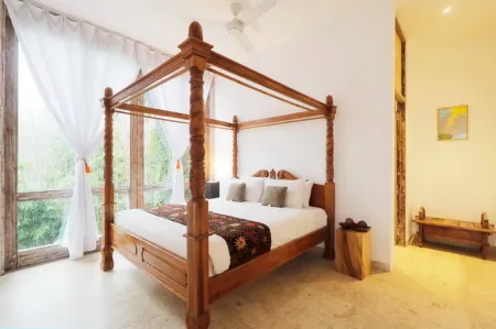 Elegant wooden four-poster canopy bed with white linens in a bright tropical bedroom, featuring sheer curtains, ceiling fan, and lush green views.