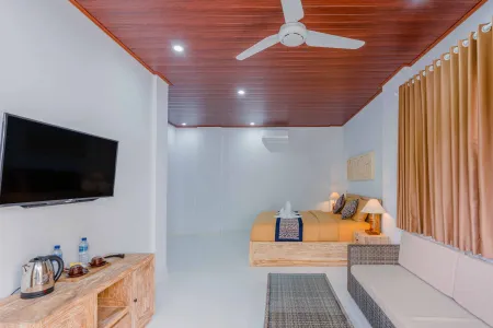 Cozy tropical bedroom with wooden accents, king bed, wall-mounted TV, ceiling fan, and beige sofa in a Bali-style villa.