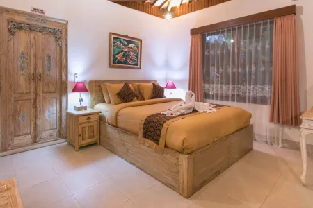 Cozy Balinese bedroom with wooden king bed, yellow linens, swan towel sculpture, koi fish painting, carved doors, and window overlooking greenery.